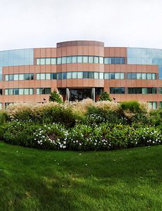 Red stone office building with many windows behind green lawn and foliage.