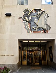 Tan colored stone building with blue, grey, red, and black mural above revolving doors next to golden 1 Rockefeller Plaza sign