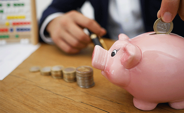 Man depositing 1 coin into piggy bank near ascending stacks of coins.