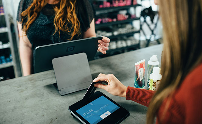 Image of woman swiping card into a sale terminal in front of another woman behind sales counter.