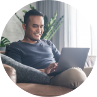 Personal Checking Smiling man in grey shirt sitting on tan couch using grey laptop.