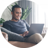 Personal Checking Smiling man in grey shirt sitting on tan couch using a laptop computer.